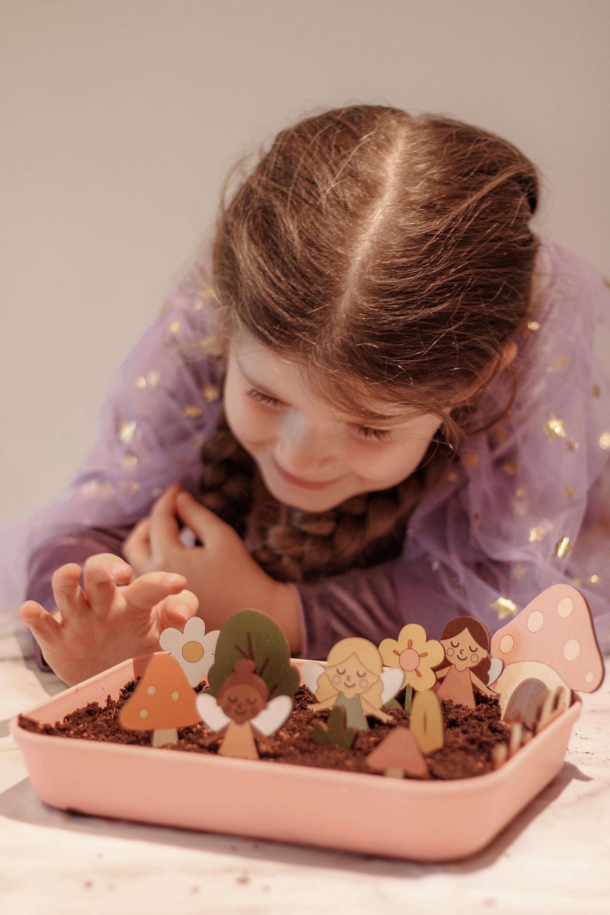 Child playing with fairy garden in a pink tray on a light surface.