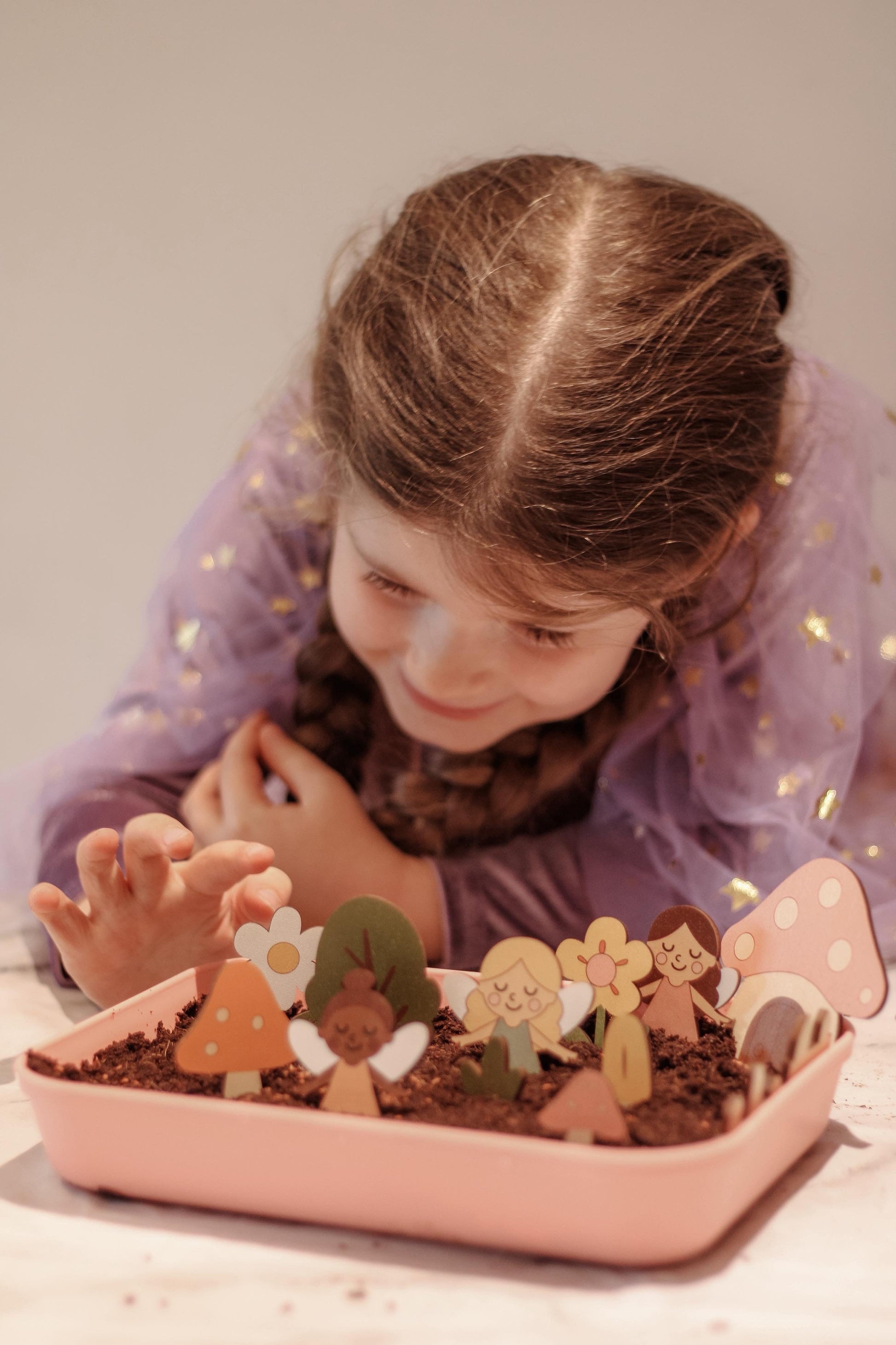 Child playing with fairy garden in a pink tray on a light surface.