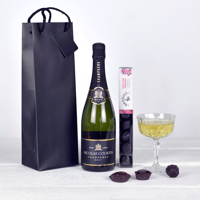 Bottle of Nicholas Colvins champagne with a gift bag, glass of champagne, and chocolate truffles on a white background