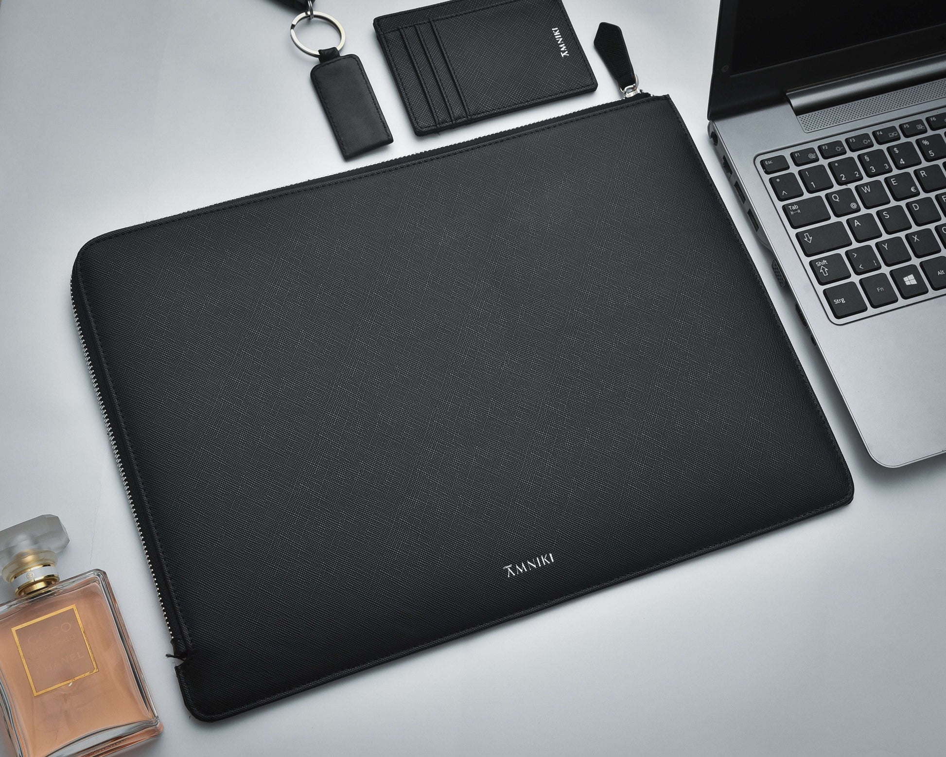 Black laptop sleeve with a laptop, perfume bottle, and wallet on a light gray background