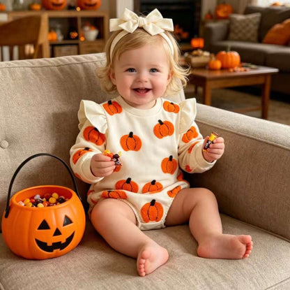 A toddler sitting on a beige couch in a cozy living room, wearing a cream Halloween romper covered in orange pumpkin patches and holding a small candy. An orange jack-o'-lantern candy bucket sits next to them.