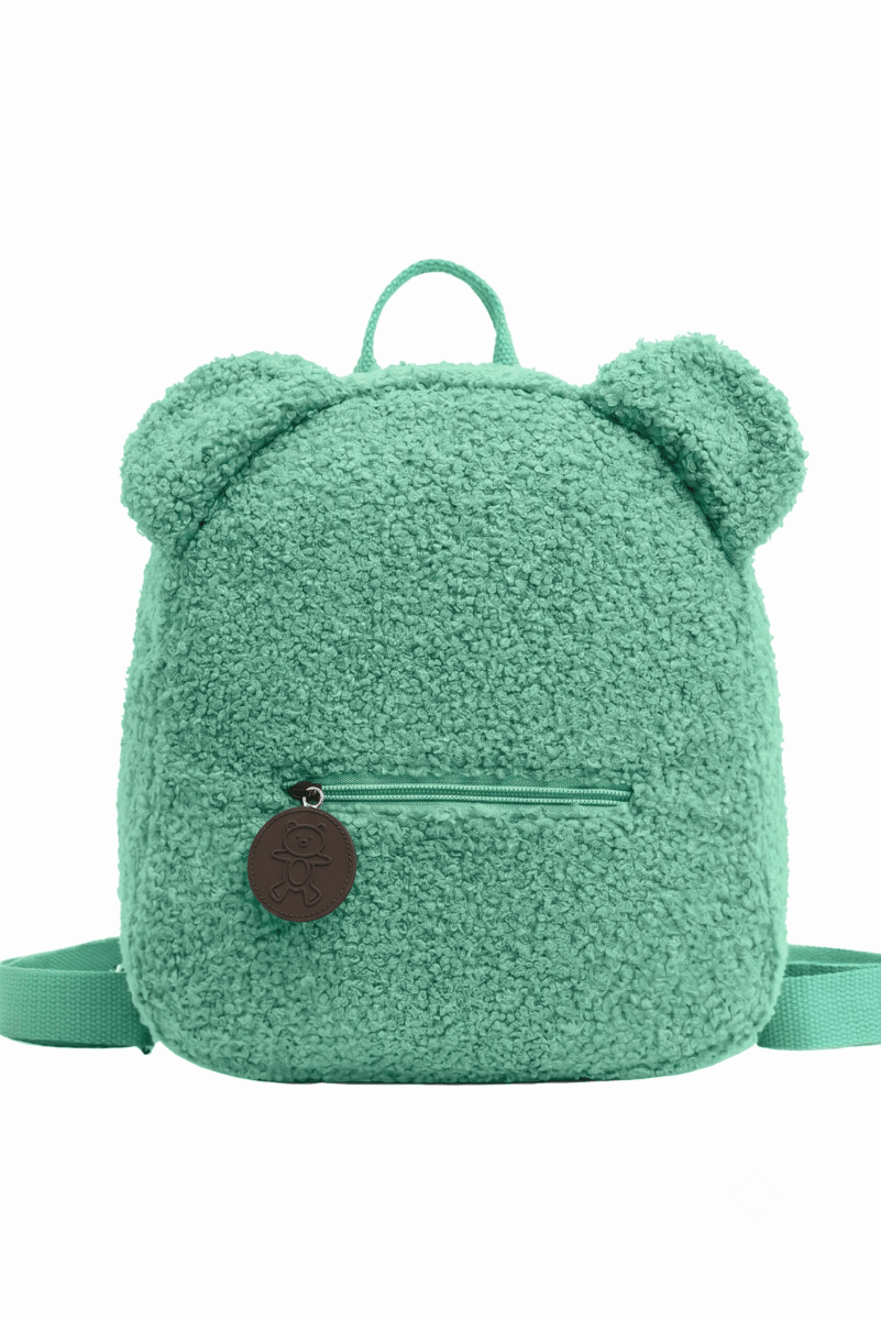 A mint green teddy fleece backpack with bear ears on top, a front zip pocket, and a round brown leather logo charm, shown against a plain white background.