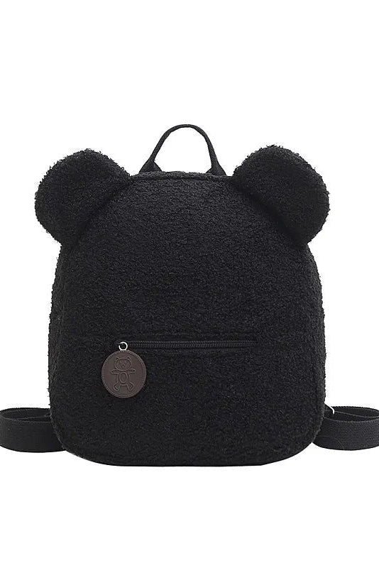 A black teddy fleece backpack with plush bear ears on the top, a top carry handle, a front zip pocket, and a round metal logo charm, shown on a plain white background.