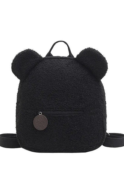 A black teddy fleece backpack with plush bear ears on the top, a top carry handle, a front zip pocket, and a round metal logo charm, shown on a plain white background.