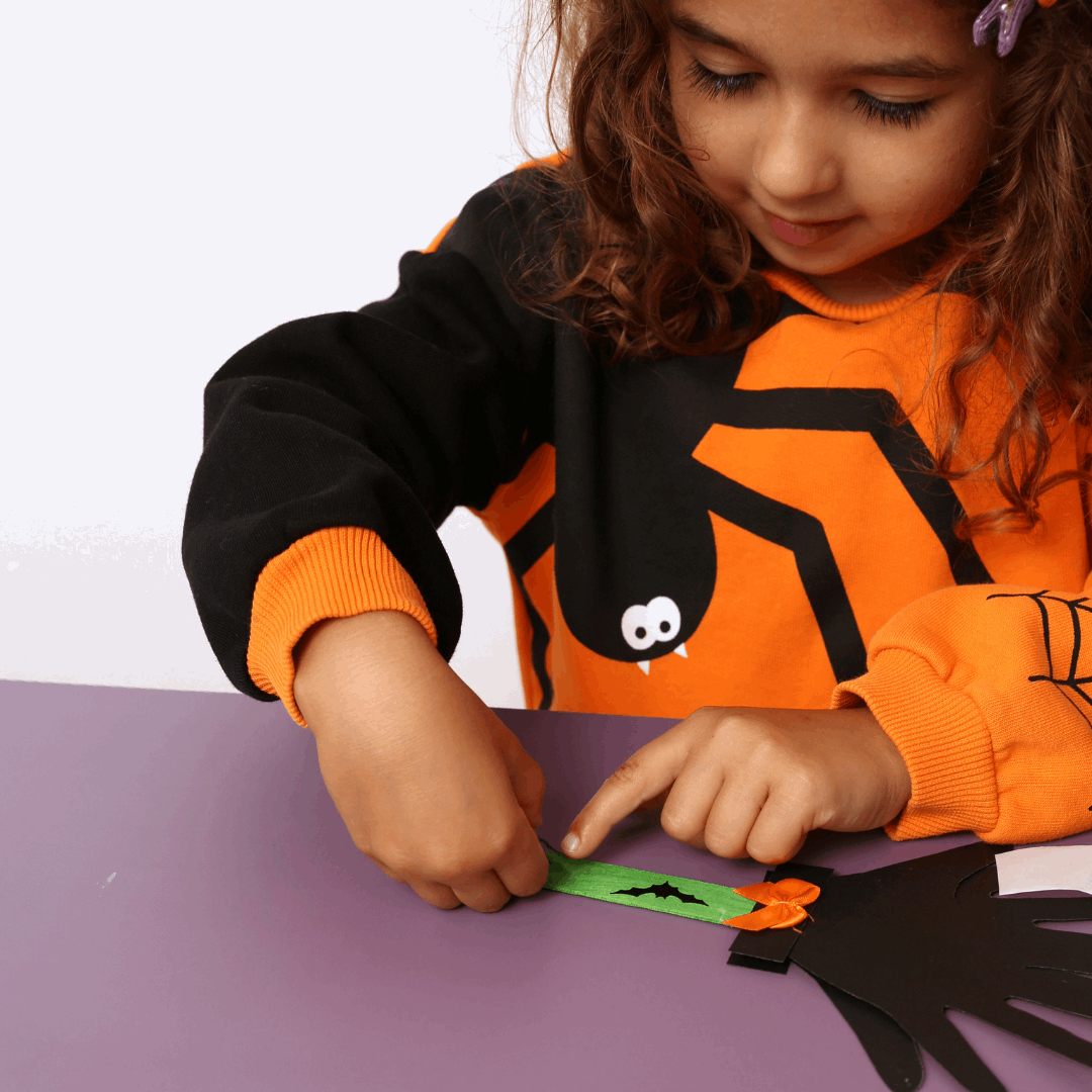 Child wearing a Halloween-themed spider shirt, sitting at a table with craft materials.