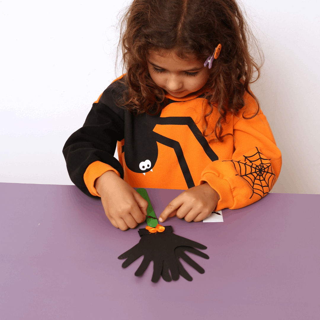 Child wearing a Halloween-themed shirt, making a craft on a purple table.