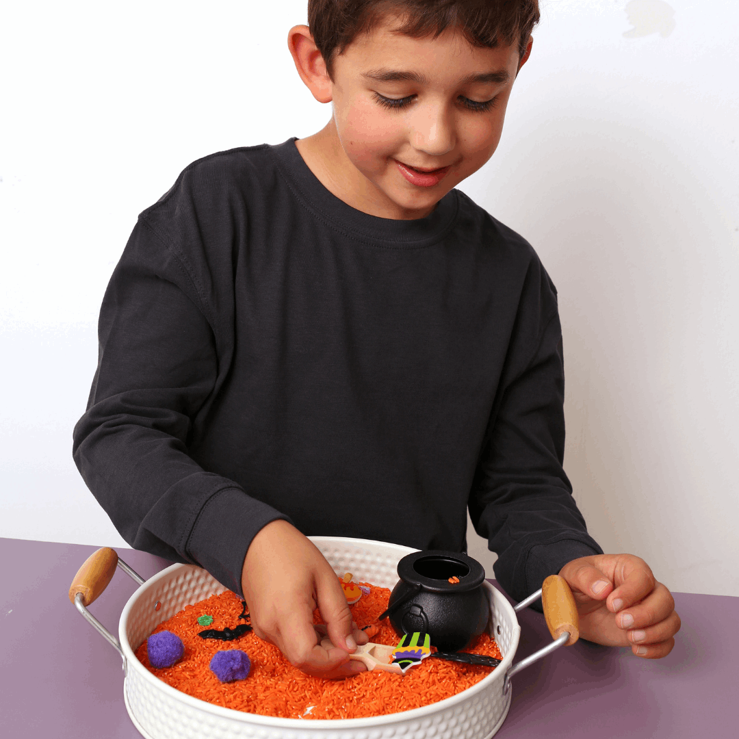 Child playing with a Halloween-themed sensory bin filled with orange sand, small toys, and a black cauldron.