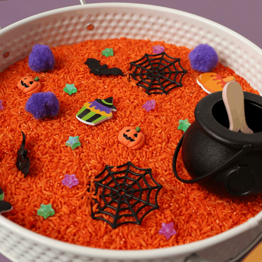 Halloween-themed sensory bin with orange rice, pumpkins, spiders, and a black pot.