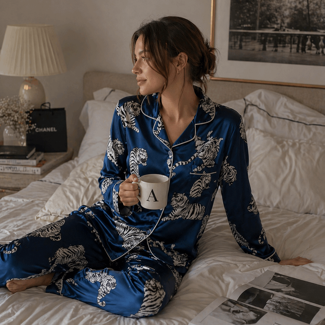 Woman in blue pajamas holding a mug on a bed with a magazine and food items.