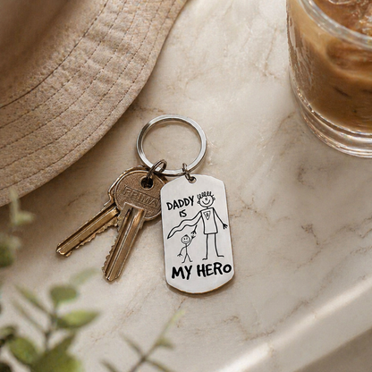 Keychain with 'Daddy is my hero' tag on a marble surface with a hat and glass of coffee.