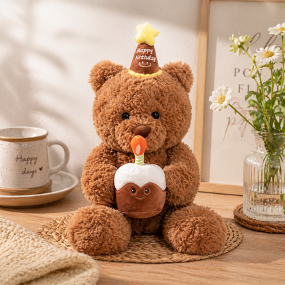 Brown teddy bear with a birthday cake and candle, sitting on a wooden table with a cup and flowers in the background.