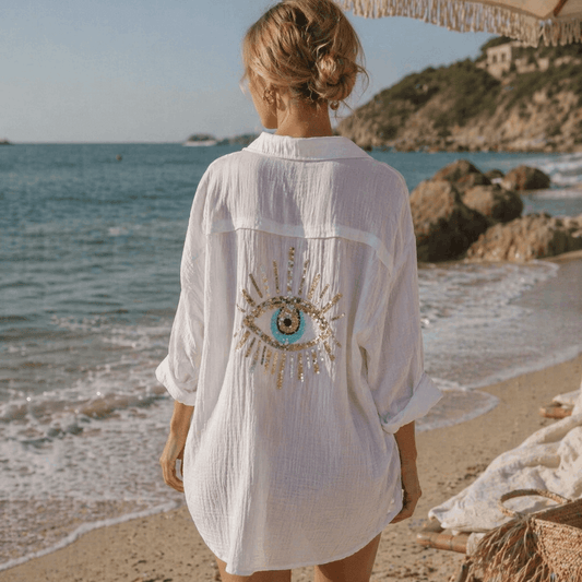 Woman in a white embroidered shirt standing on a beach with ocean and rocks in the background