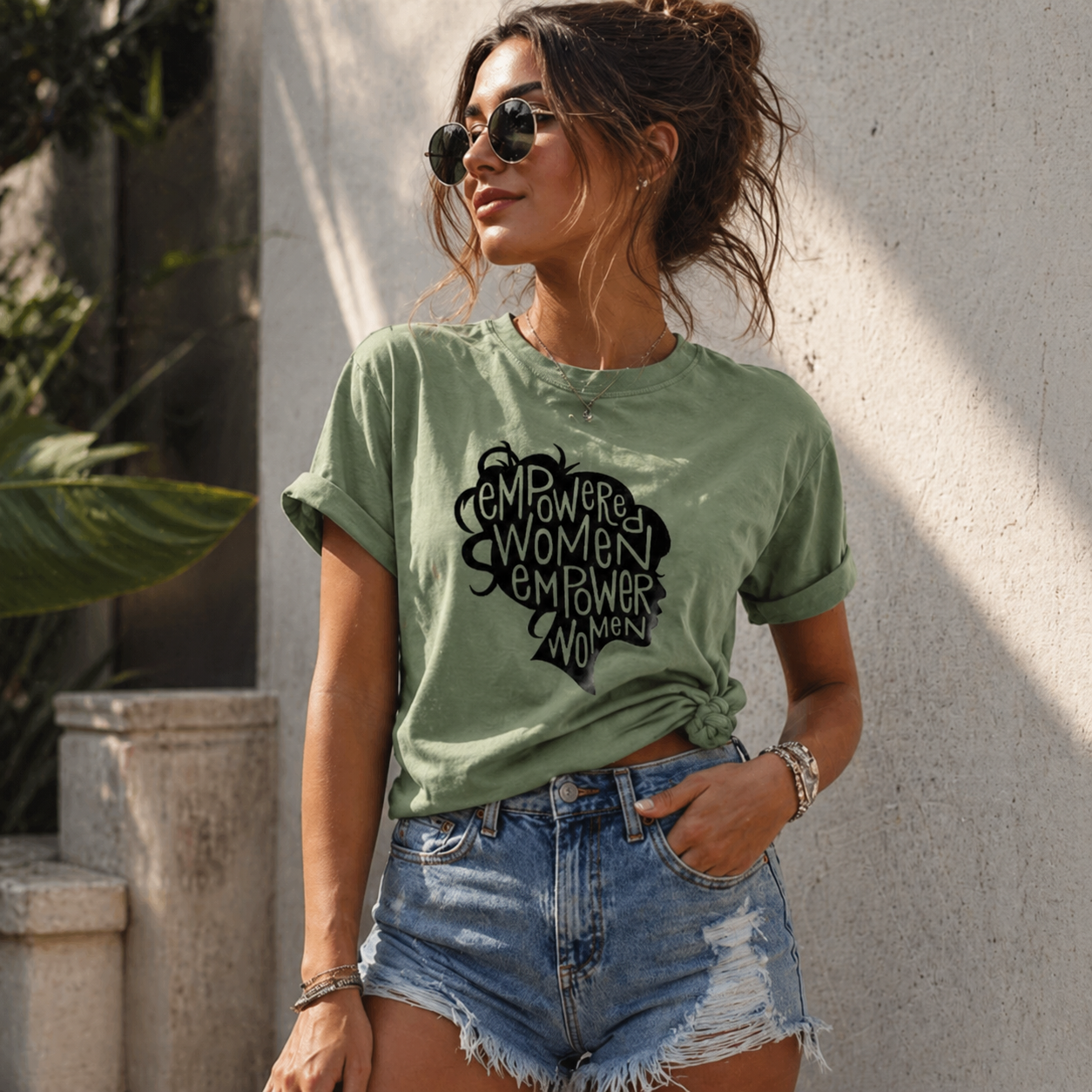 Woman wearing a green t-shirt with a black graphic and text, standing outdoors against a light-colored wall.