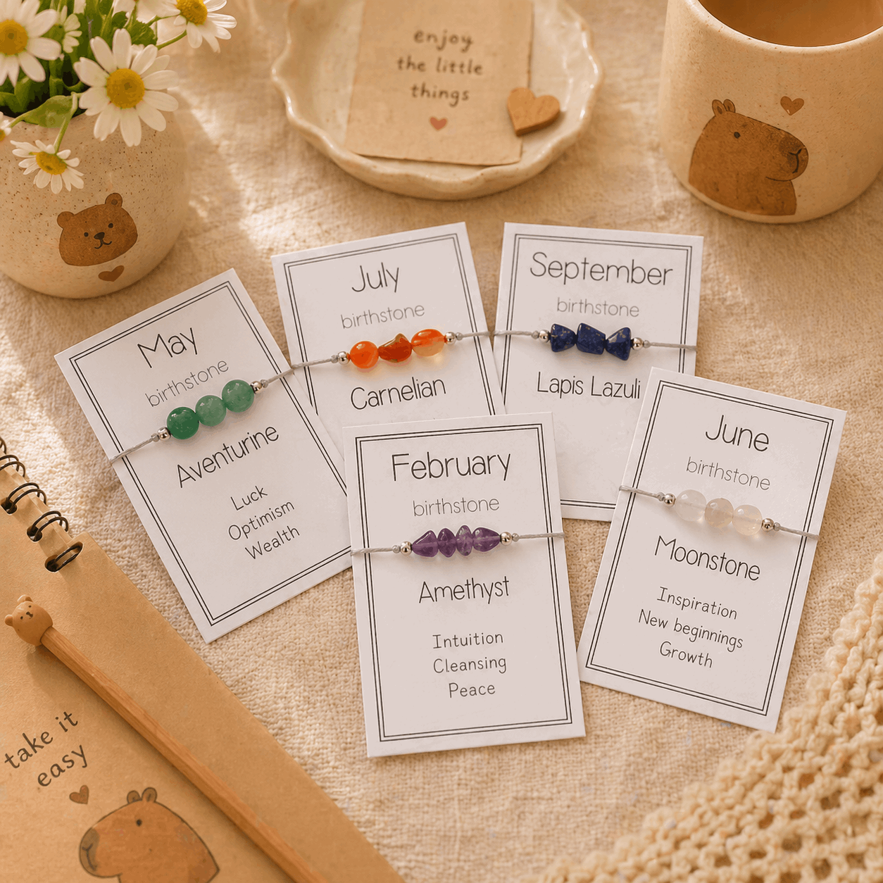 Set of birthstone bracelets on cards with descriptive text, placed on a textured surface with a cup and flowers in the background.