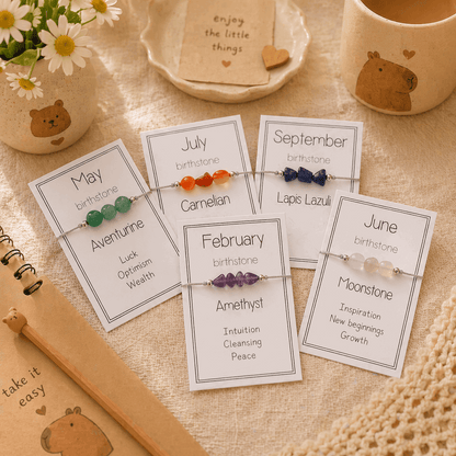 Set of birthstone bracelets on cards with descriptive text, placed on a textured surface with a cup and flowers in the background.