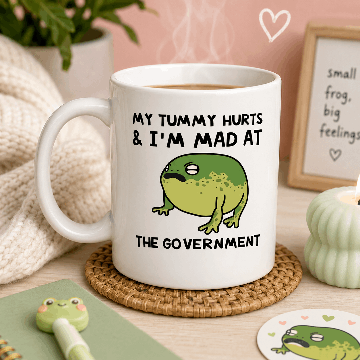 White mug with a frog illustration and text on a table with a heart-shaped frame in the background