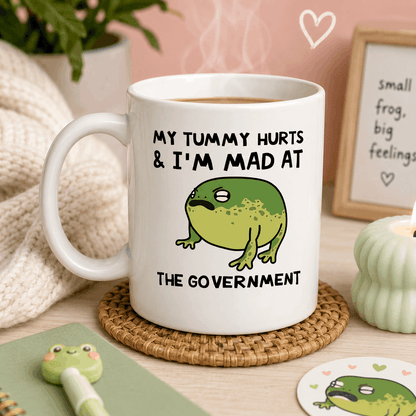 White mug with a frog illustration and text on a table with a heart-shaped frame in the background