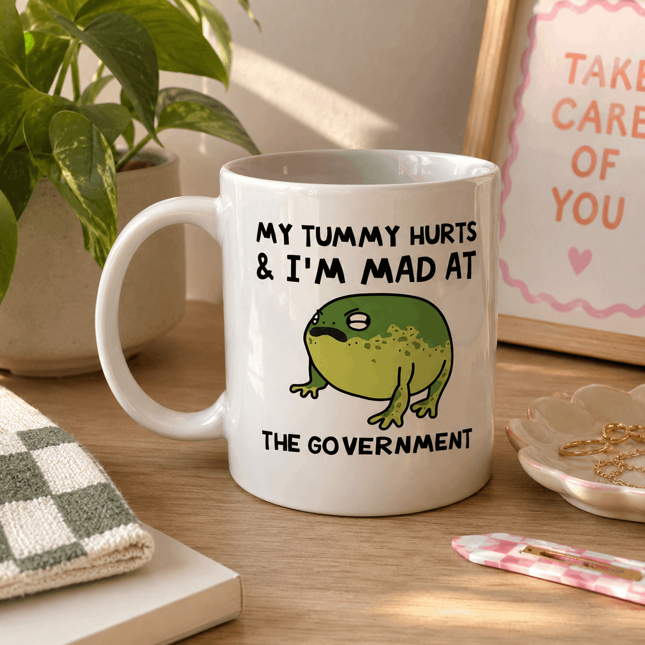 White mug with a frog graphic and text on a wooden surface with a plant and framed picture in the background.
