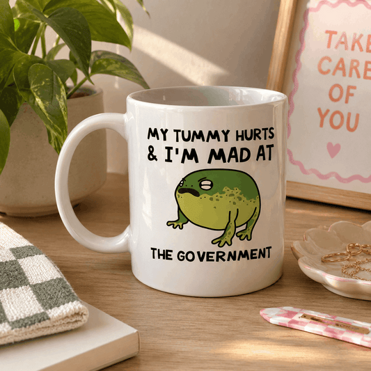 White mug with a frog graphic and text on a wooden surface with a plant and framed picture in the background.