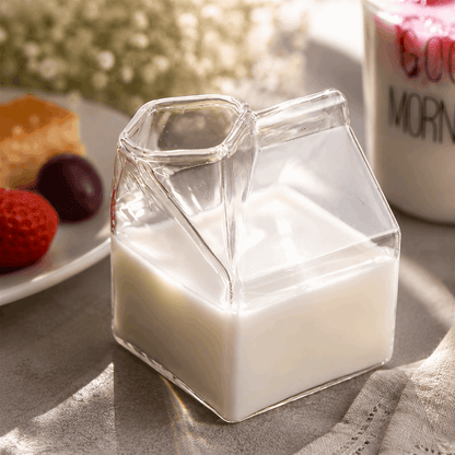 Glass milk carton filled with milk on a table with a plate of fruit and a mug in the background.