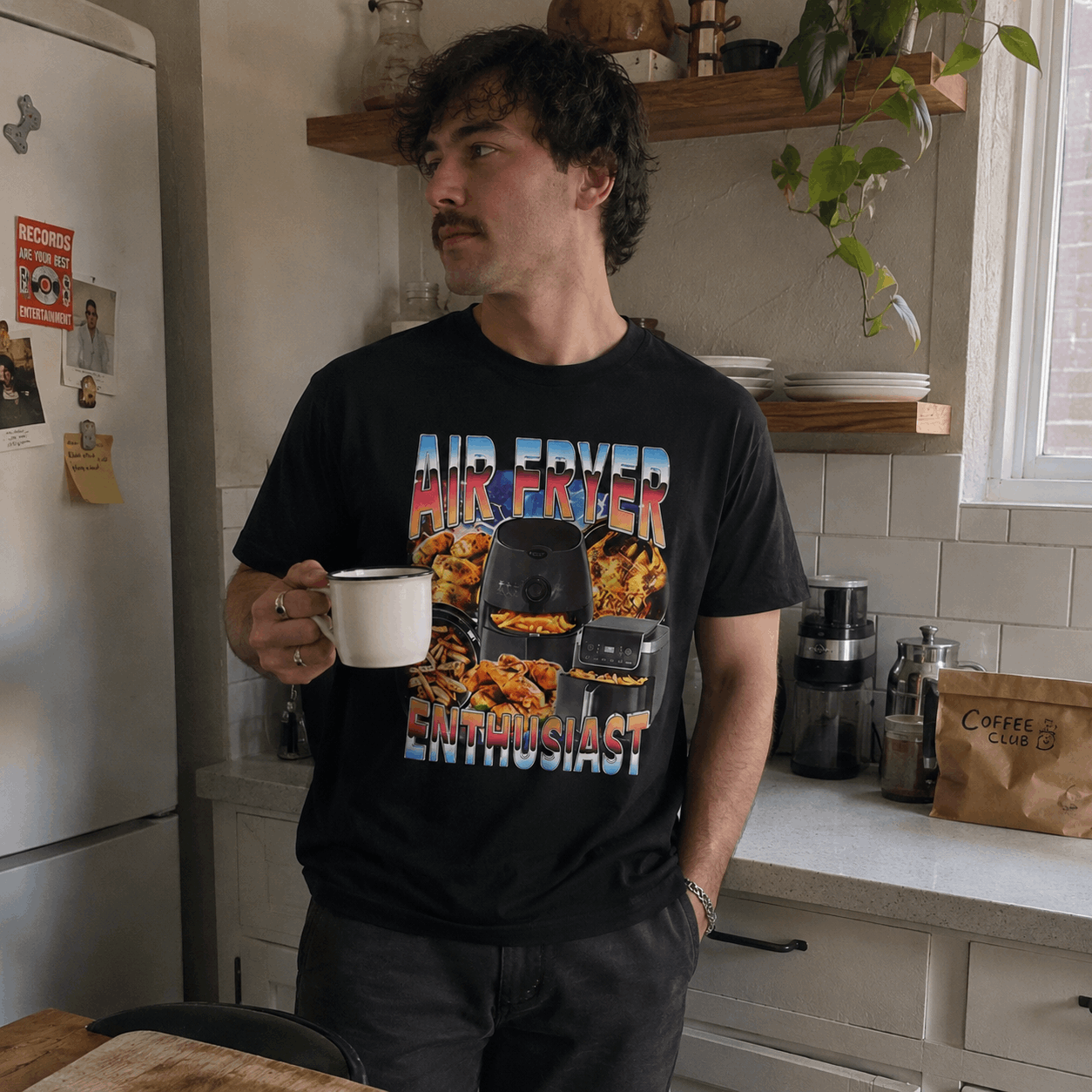 Man in a kitchen holding a mug, wearing a black t-shirt with colorful graphics and text.