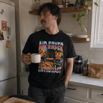 Man in a kitchen holding a mug, wearing a black t-shirt with colorful graphics and text.