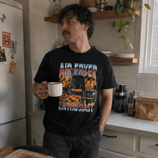 Man in a kitchen holding a mug, wearing a black t-shirt with colorful graphics and text.
