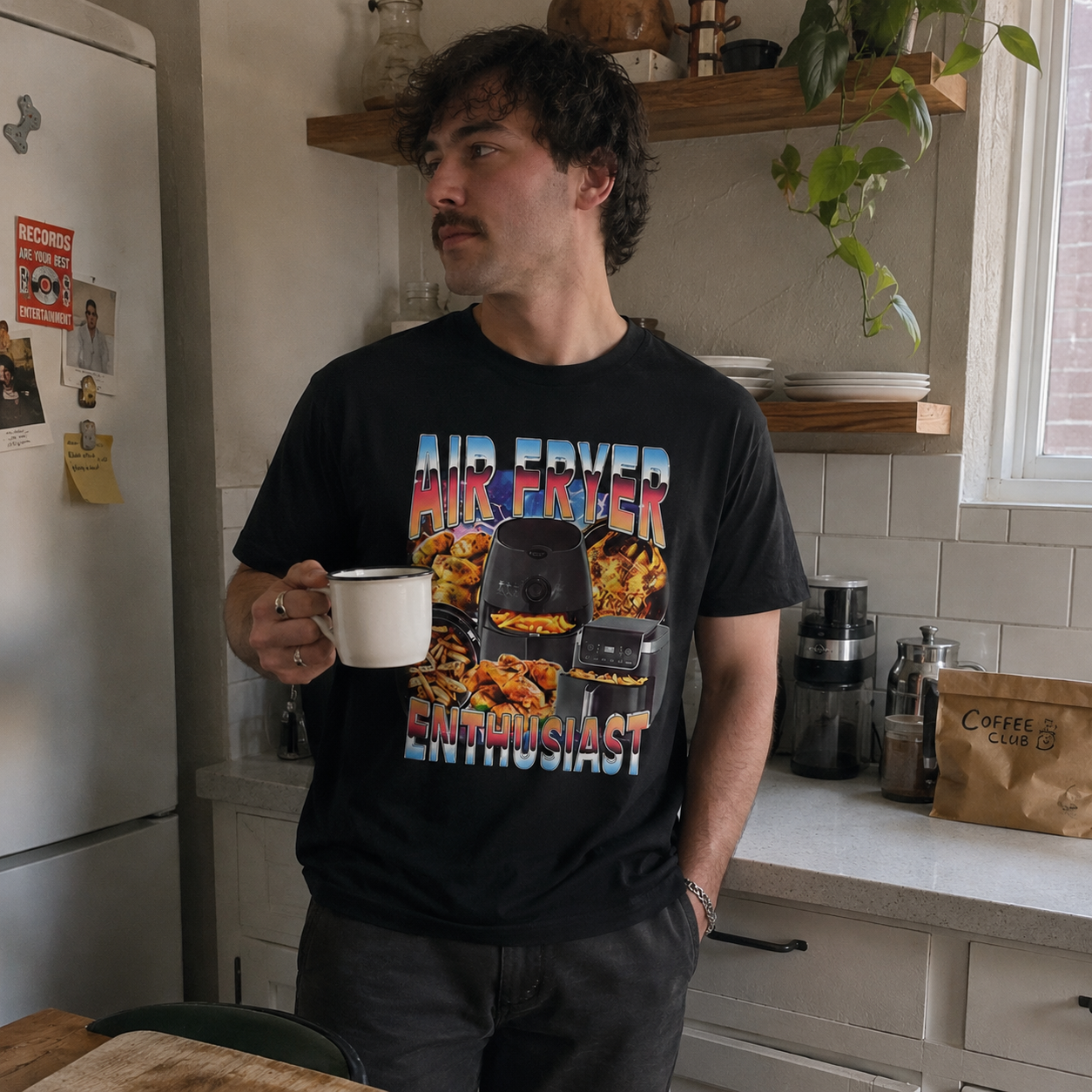 Man in a kitchen holding a mug, wearing a black t-shirt with colorful graphics and text.