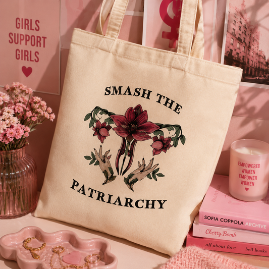 Beige tote bag with 'Smash the Patriarchy' design on a pink background with books and flowers.