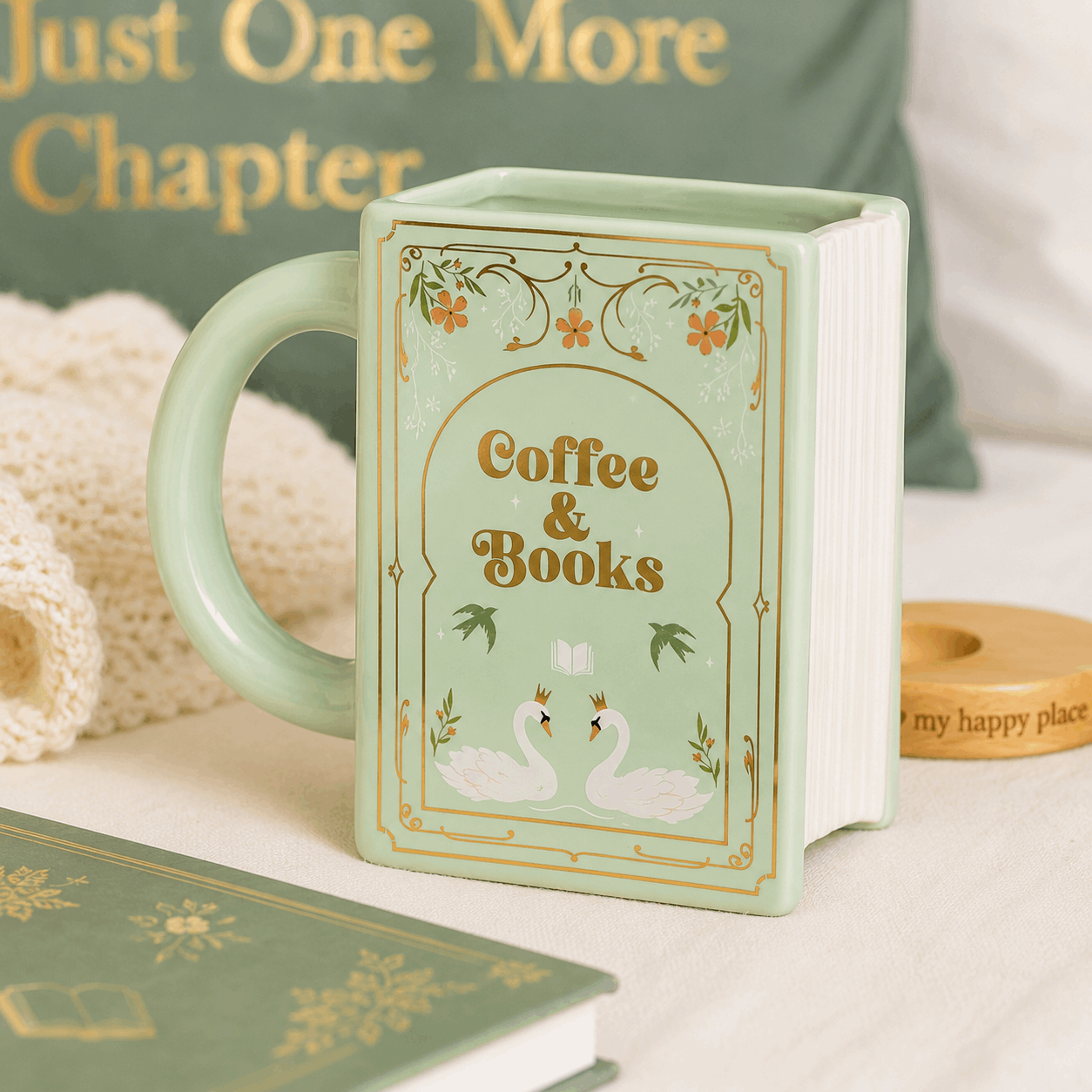 Mint green mug designed like a book with 'Coffee & Books' text and swan illustration, placed on a surface with books and a cozy blanket.