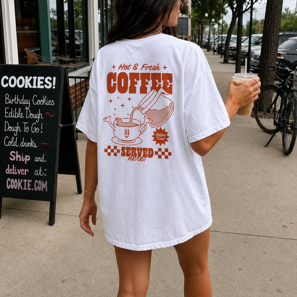 Person wearing a white t-shirt with coffee-themed design, holding a drink on a sidewalk.