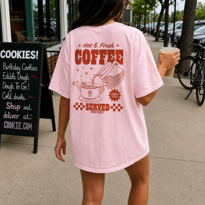 Person wearing a pink t-shirt with coffee-themed design, holding a drink on a sidewalk.
