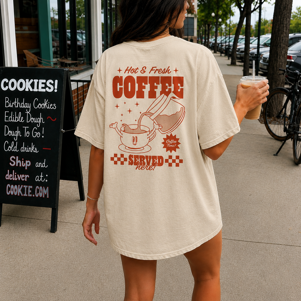 Person wearing a t-shirt with coffee-themed design, holding a drink, walking outdoors.