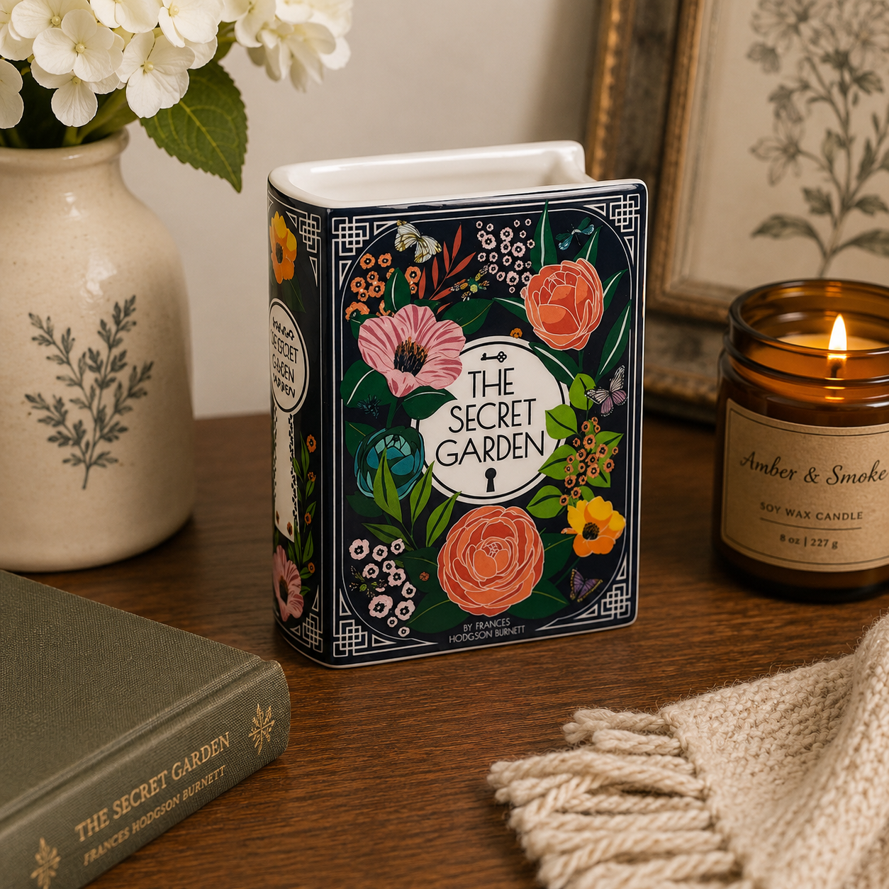 Decorative book-shaped container with floral design on a wooden surface next to a candle and vase.
