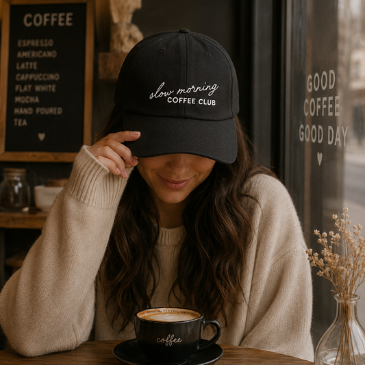 Woman wearing a black cap with 'slow morning COFFEE CLUB' text, sitting at a table with a coffee cup in a cozy setting.