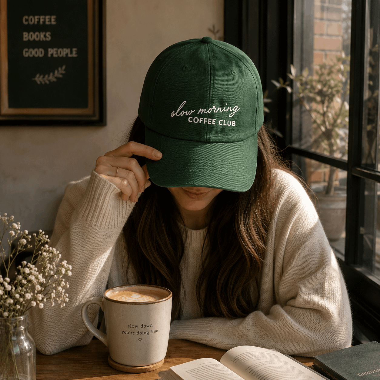 Person wearing a green cap with 'slow morning COFFEE CLUB' text, sitting at a table with a coffee mug and book.