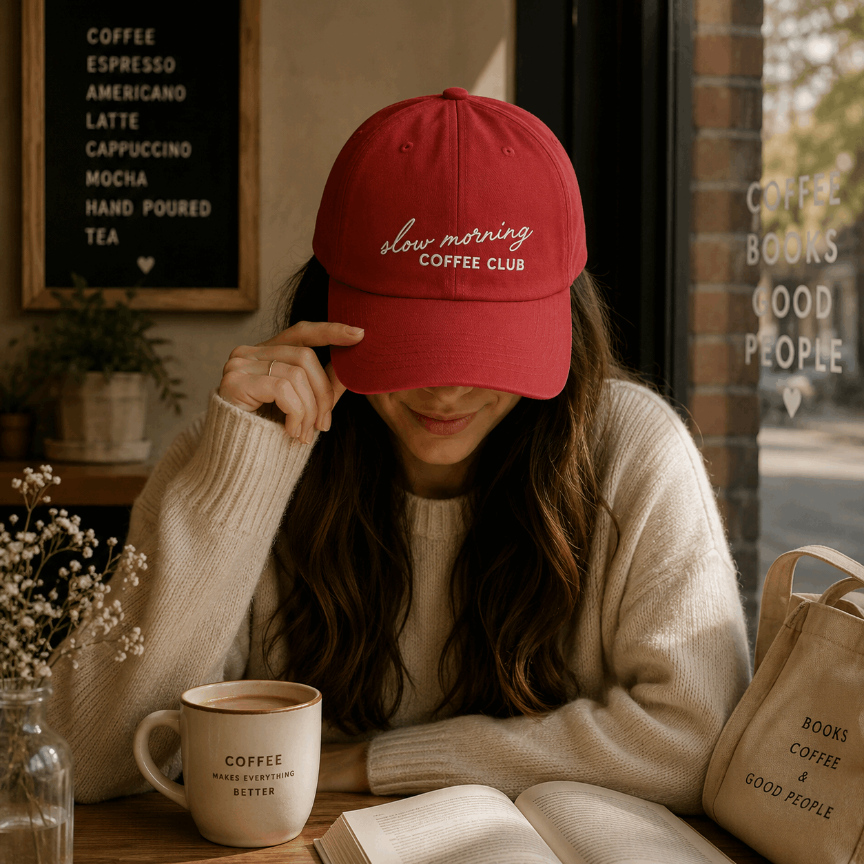 Woman wearing a red cap with 'Coffee Club' text, sitting at a table with a coffee cup and book in a cozy setting.