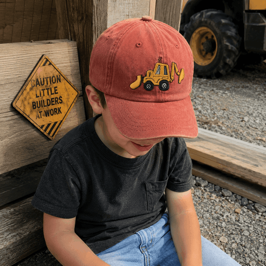 Child wearing a red cap with a bulldozer emblem, sitting next to a wooden block and warning sign.