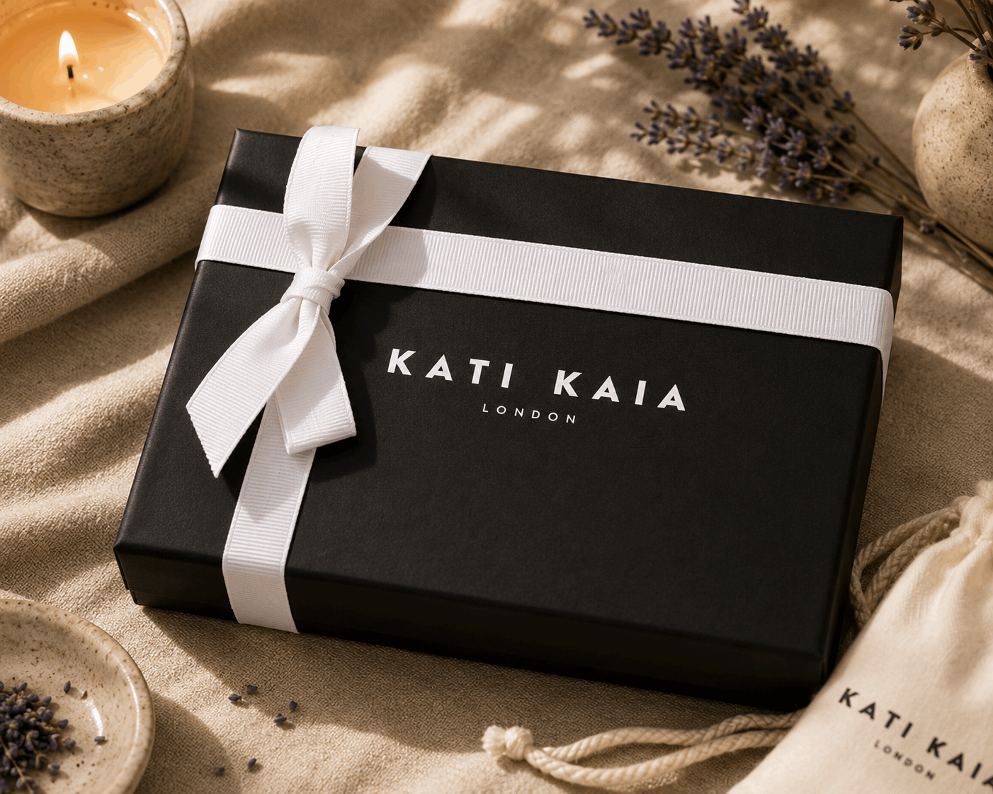 Black gift box with white ribbon and 'Kati Kaia London' branding on a textured surface with lavender and a candle.
