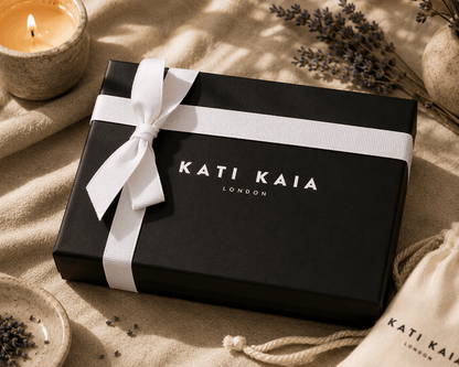 Black gift box with white ribbon and 'Kati Kaia London' branding on a textured surface with lavender and a candle.