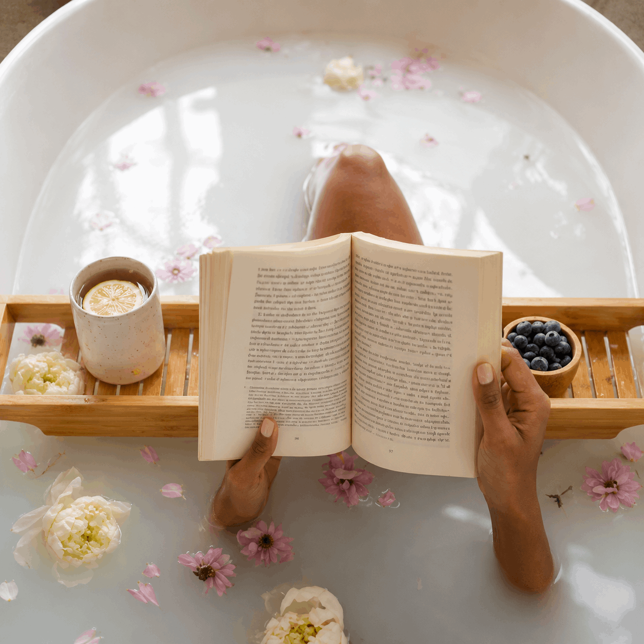 Person reading a book in a bathtub filled with water and flowers, with a tray of items including a cup and bowl.