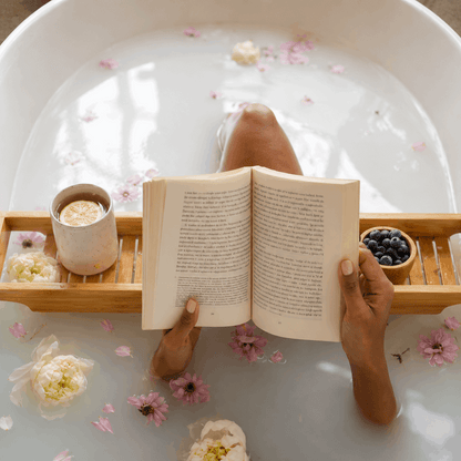 Person reading a book in a bathtub filled with water and flowers, with a tray of items including a cup and bowl.