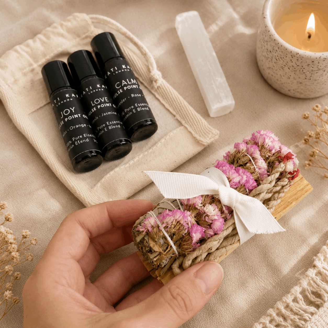 Three black rollerball bottles labeled 'Joy', 'Love', and 'Calm' on a beige fabric background with a hand holding a small woven basket with pink flowers.