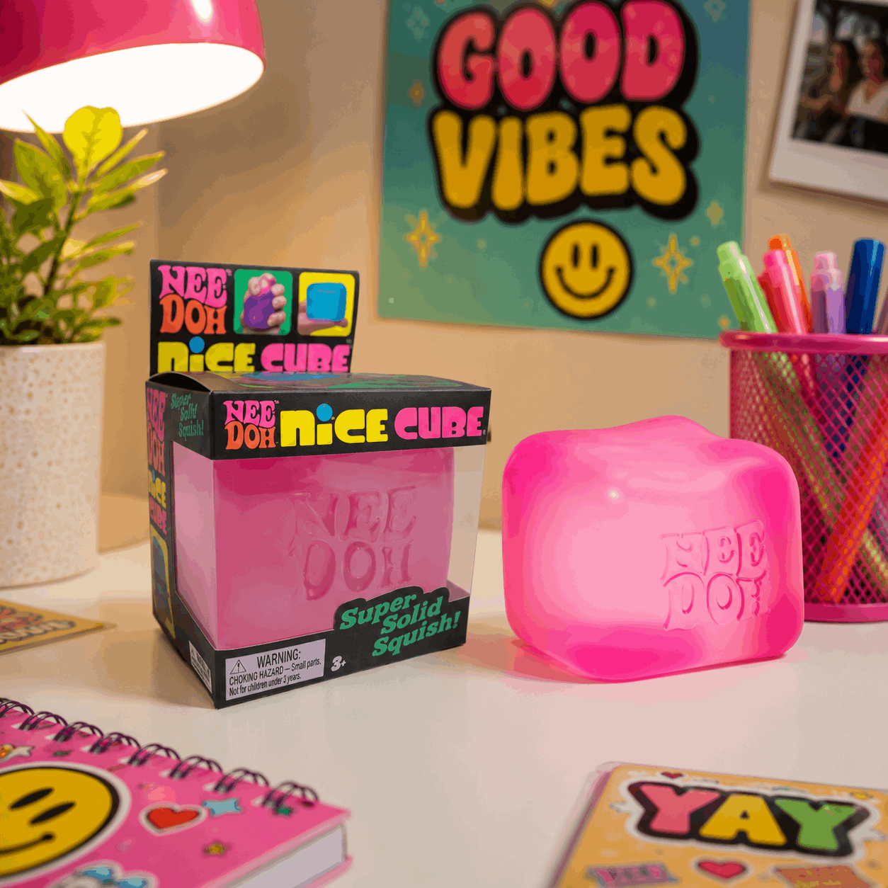Pink 'Nee Doh Nice Cube' on a desk with 'Good Vibes' poster and stationery items.