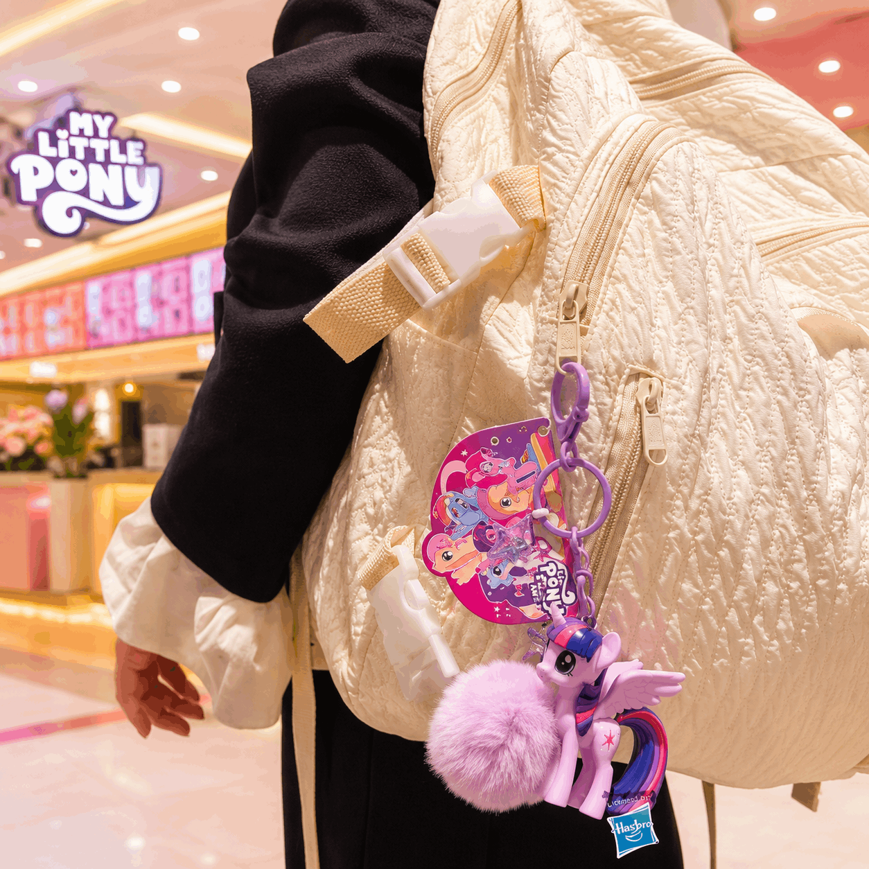 Person wearing a cream jacket with a colorful keychain featuring 'My Little Pony' characters in a mall setting.