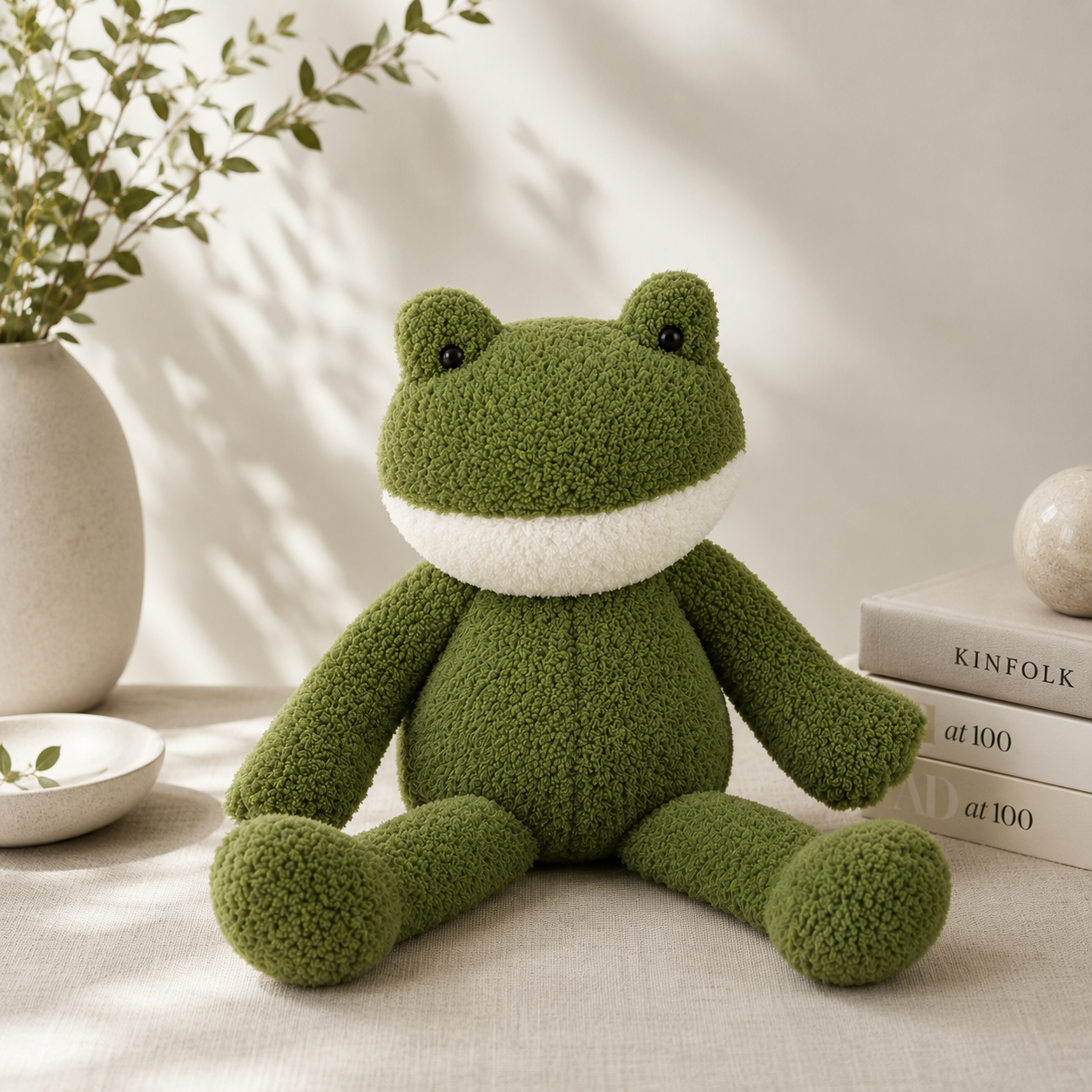 Green plush frog toy on a light surface with books and a plant in the background