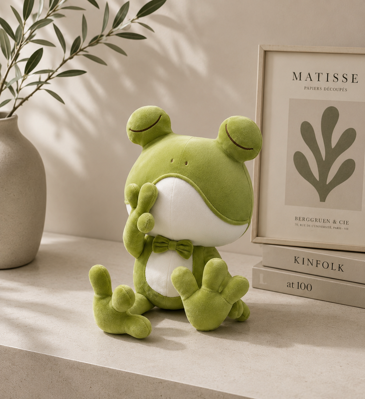Green plush frog toy on a surface with a plant and framed picture in the background