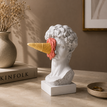 Statue head with ice cream cone on a table next to a book and vase.