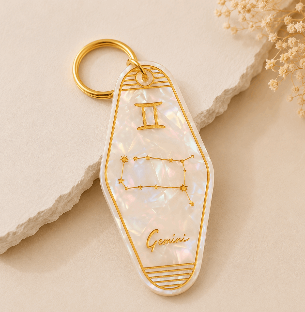 Keychain with Gemini zodiac design on a beige stone surface with dried flowers.