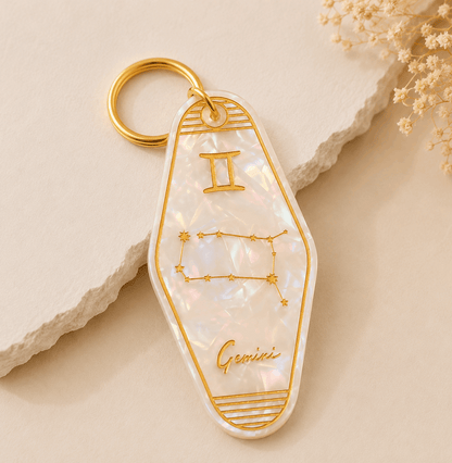 Keychain with Gemini zodiac design on a beige stone surface with dried flowers.